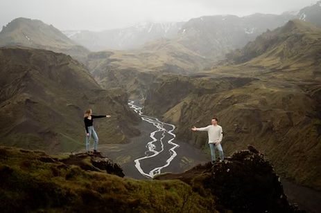 engaged couple stretching arms at each other on a cliff in Thorsmork Iceland.