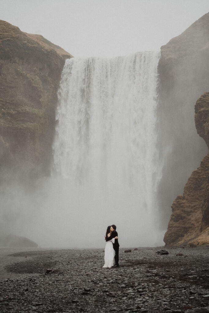 A couple embraces in front of a large, misty waterfall. The rocky ground contrasts with the bride's white dress, creating a romantic scene during diamond beach elopement