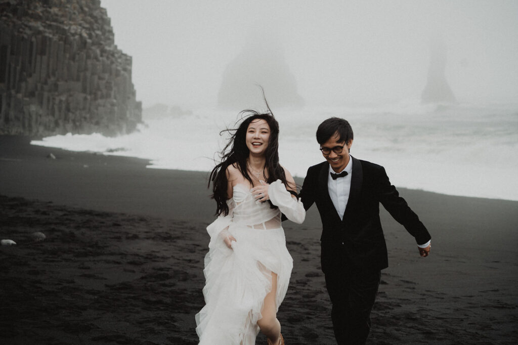 bride and groom run on black sand beach during iceland adventure wedding.