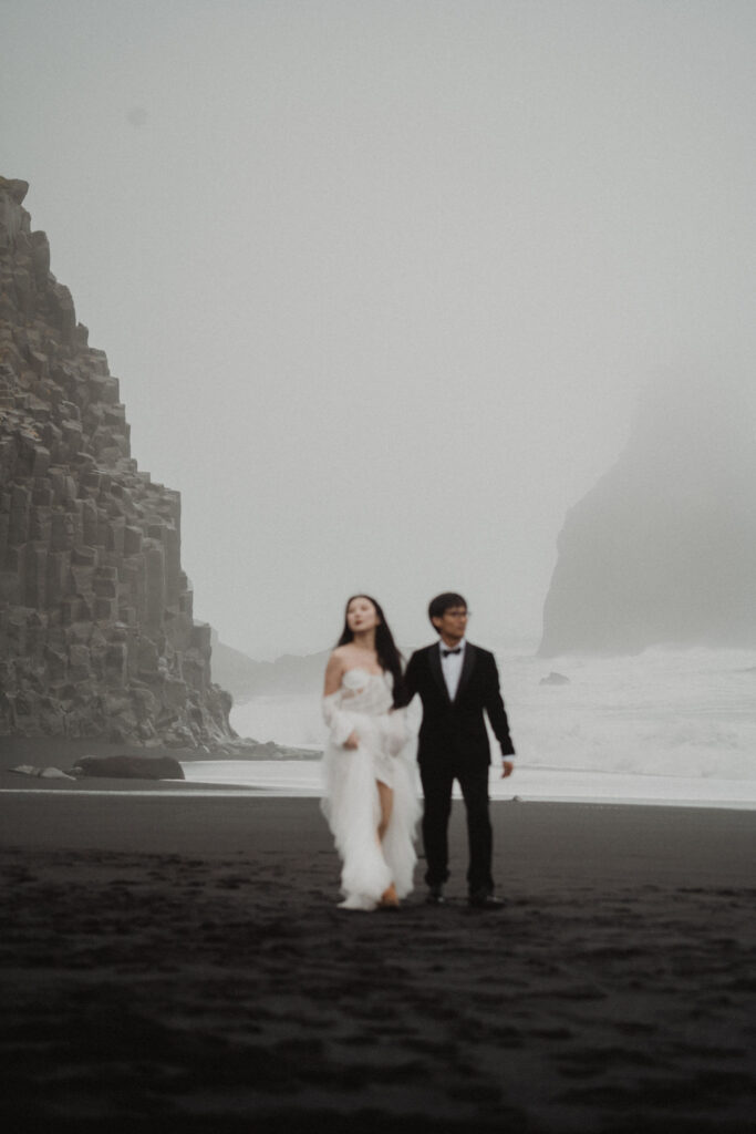bride and groom runs on a foggy black sand beach with rocky cliffs during diamond beach elopement