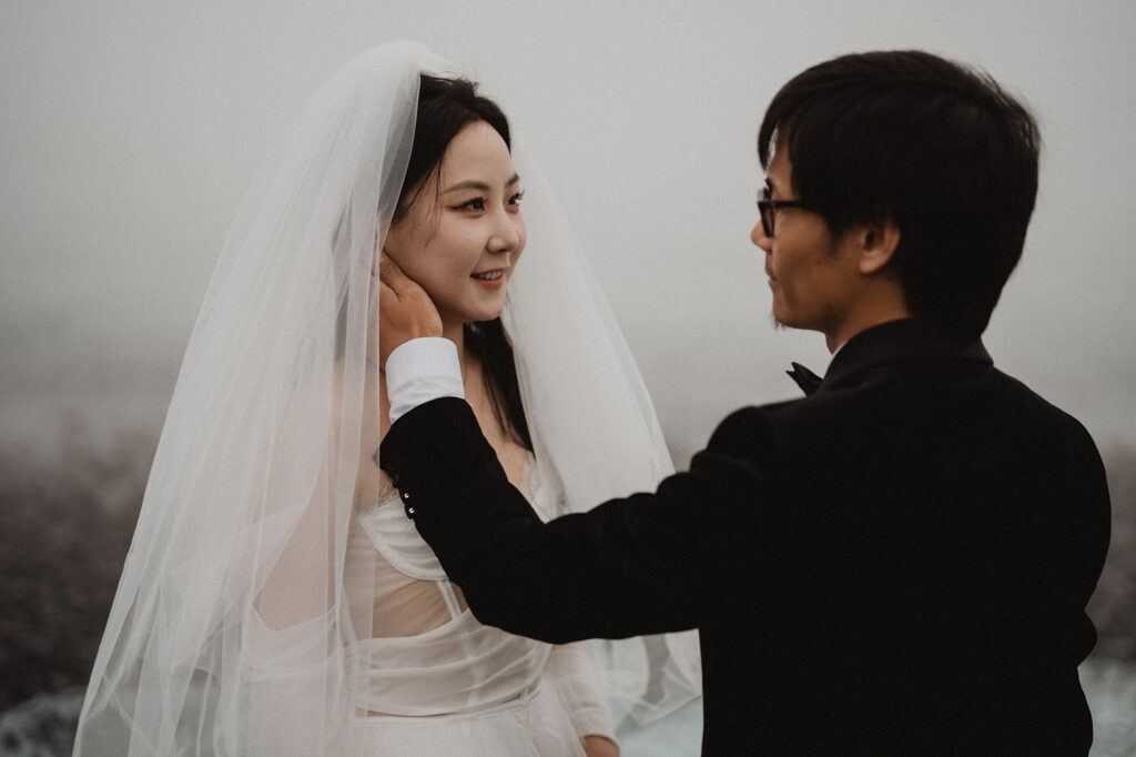 Bride in white smiling at groom in black suit, gently touching her face. Misty, neutral background at their diamond beach elopement.