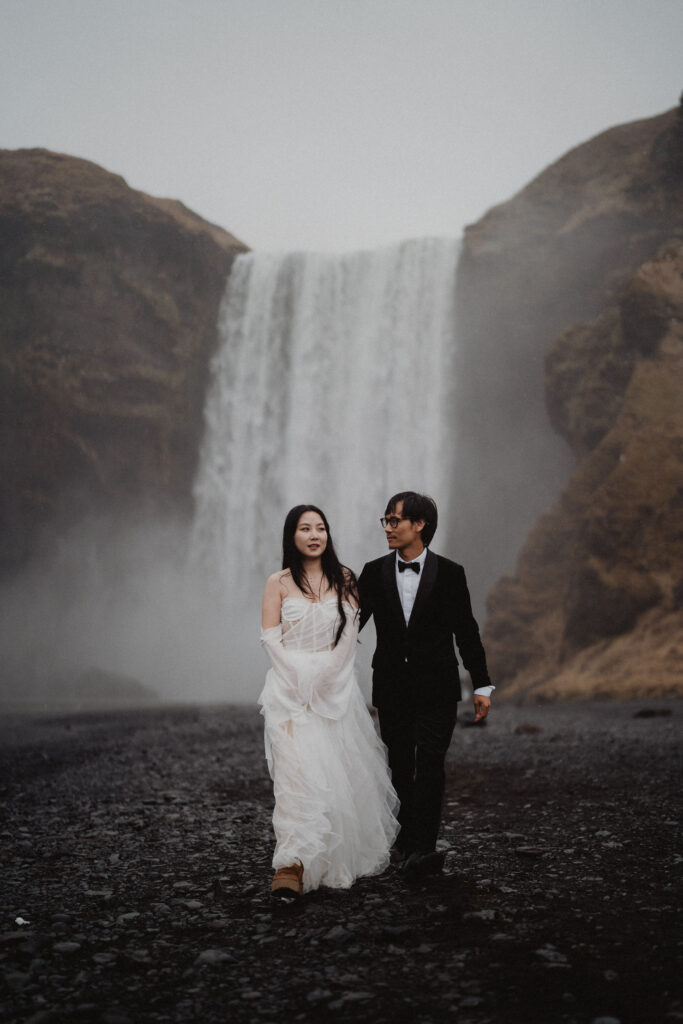 bride and groom walking in front of a majestic waterfall, set against misty, rocky terrain. The mood is romantic and serene for their diamond beach elopement