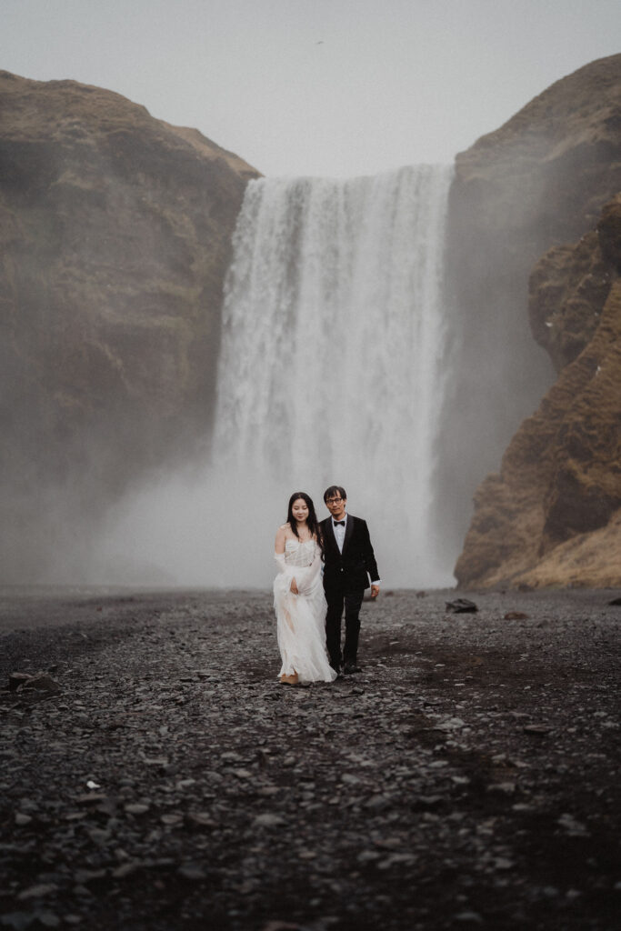 bride and groom walk on rocky ground with a cascading waterfall in the misty background during diamond beach elopement
