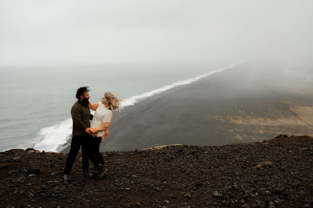 A couple in cozy sweaters hug and look at each other on a cliff overlooking the black sand beach during honeymoon photo shoot with Iceland wedding photographer.