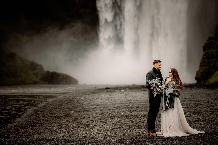 Bride and groom hold flowers, smiling in front of a misty waterfall on rocky ground, set against a moody, natural backdrop.