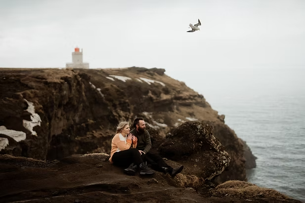 A couple sits on the cliff edge with a lighthouse int he background during honeymoon pictures with Iceland Wedding Photographer.