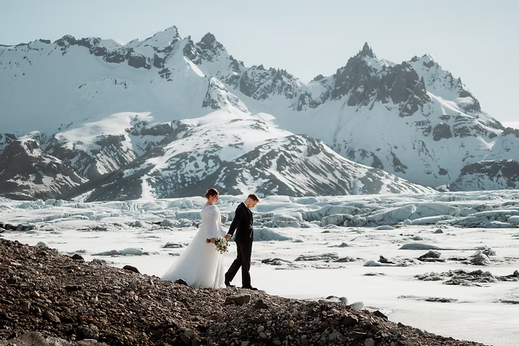 Bride in white gown and groom in black suit walk hand in hand on rocky Iceland terrain with snowy mountains and glaciers in the background while they get married