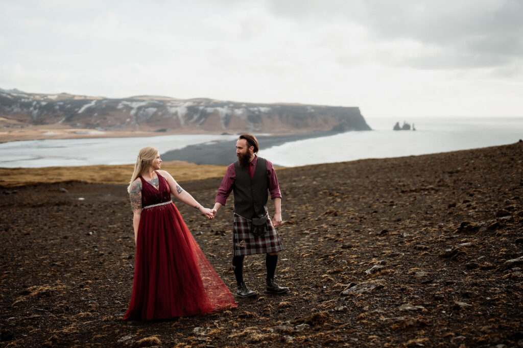 During honeymoon photo shoot with Iceland Wedding Photographer, a couple in red stand hand in hand with the sea and mountains in the background.