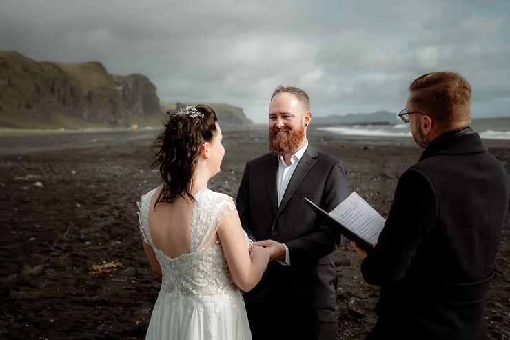 how to get married in iceland: Couple holding hands on rocky beach during wedding ceremony, with officiant reading. Overcast sky, cliffs, and ocean in background.