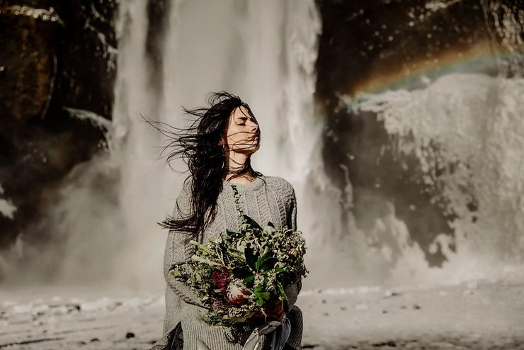 how to get married in iceland: A woman in a sweater holds an elopement bouquet, standing by a waterfall with a faint rainbow in the mist. Her hair flows in the wind, exuding calm.