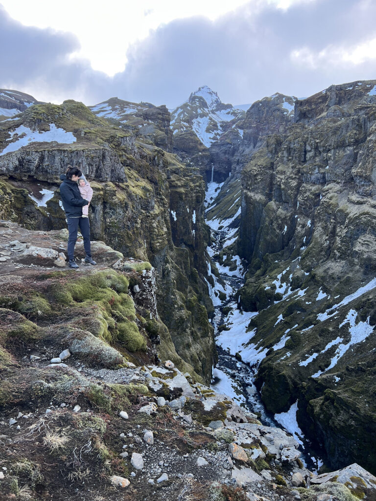 Iceland Wedding Photographer takes photos during a hike in Iceland with her family.  She stands on the edge of the valley, snowy mountains in the background, holding her small daughter.