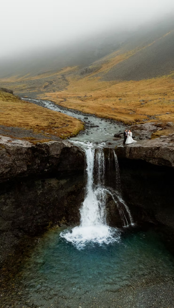 after learning how to get married in iceland, bride and groom standing near a waterfall, surrounded by misty mountains and a winding stream.
