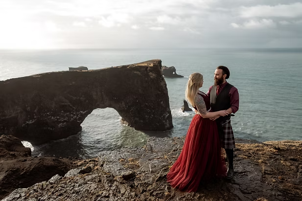 A couple enbraces and smiles at each other, with cliffs over the sea in the backgrouAnd, during Honeymoon Photo shoot with Iceland Wedding Photographer.