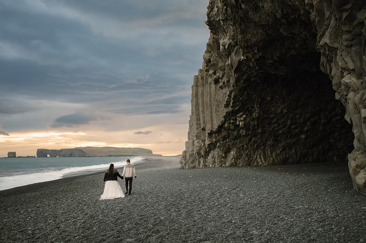 Couple walks on a black sand beach in front of a large cave. Overcast sky and distant sea cliffs in iceland during wedding