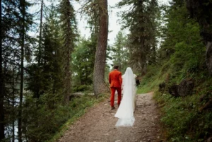 bride and groom walk up dirt trail surrounded by pine trees during dolomites elopement in italy