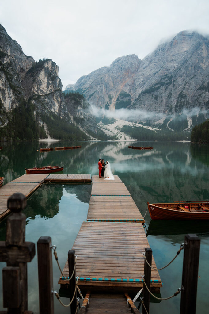 Bride and groom stand on a dock at Lago di Braies, surrounded by misty mountains during wedding ceremony. 