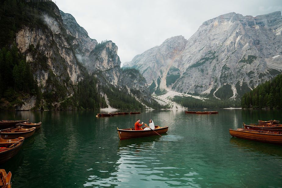 bride and groom in a wooden boat on Lago di Braies, surrounded by mountains and trees. Other boats are visible; cloudy sky above the dolomites in italy