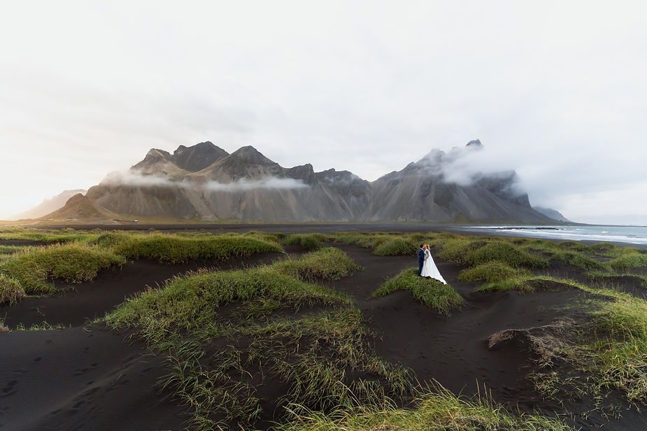 Secret Iceland Elopement Spots: Bride and groom stand amid lush green grass and black sand with misty mountains in the background, creating a serene, romantic scene.
