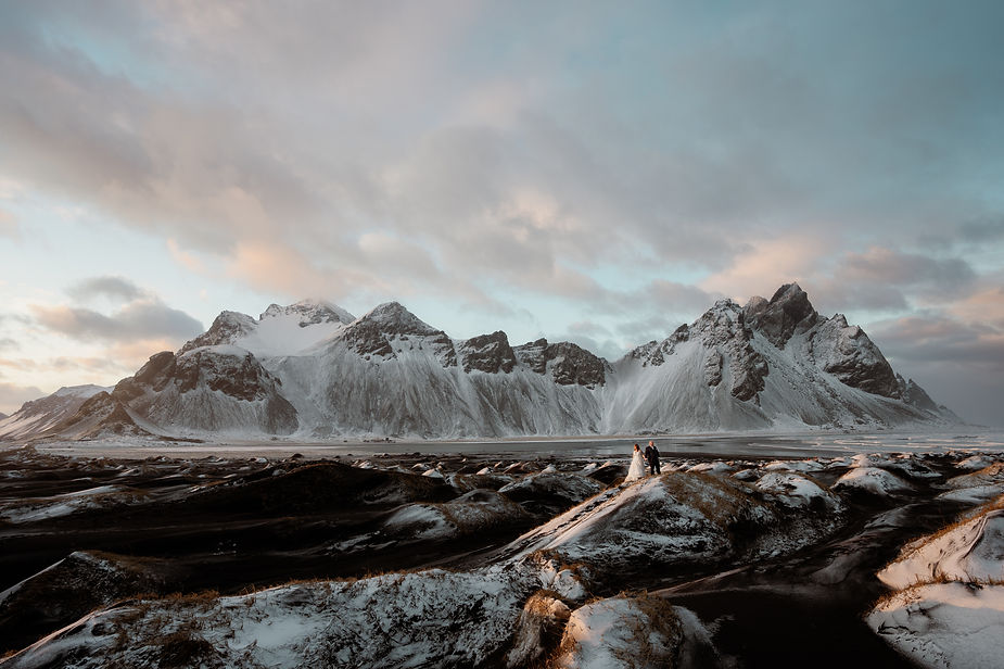 Secret Iceland Elopement Spots: Bride and groom stand on snowy terrain with majestic mountains in the background under a cloudy sky, creating a serene, romantic scene.