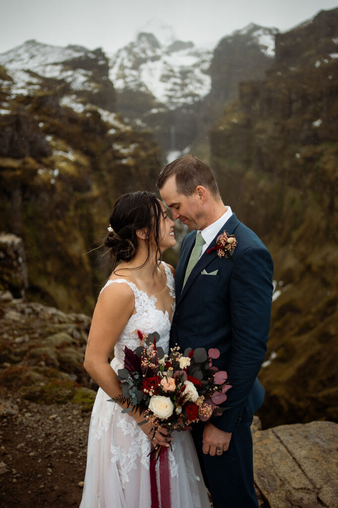 secret iceland elopement spots: Bride and groom embrace, smiling against a backdrop of rugged, snow-dusted mountains. Bride holds a colorful bouquet. Romantic mood.
