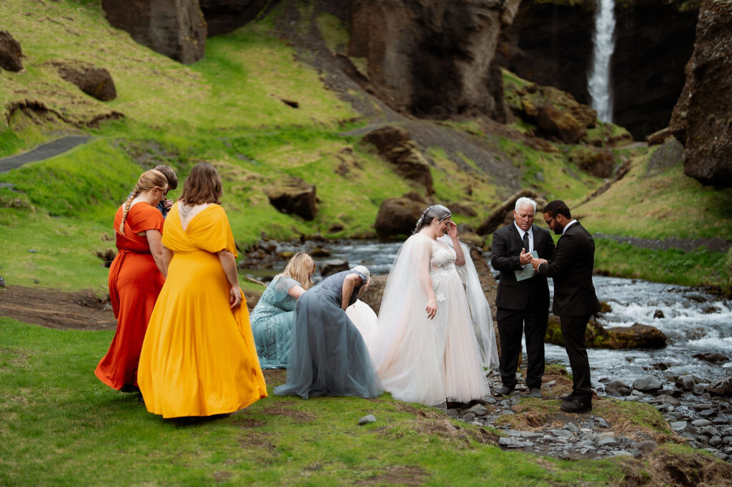 How to Include Your Family When You Elope: Bride and groom stand by a stream, surrounded by bridesmaids in colorful dresses. A waterfall and green hills in the background. Outdoor wedding.