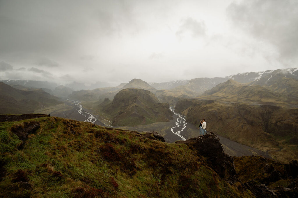 Thórsmörk: The Valley of Thor | How To Plan An Elopement Here:  couple stands on a rocky cliff overlooking a vast, green valley with winding rivers and cloudy skies.
