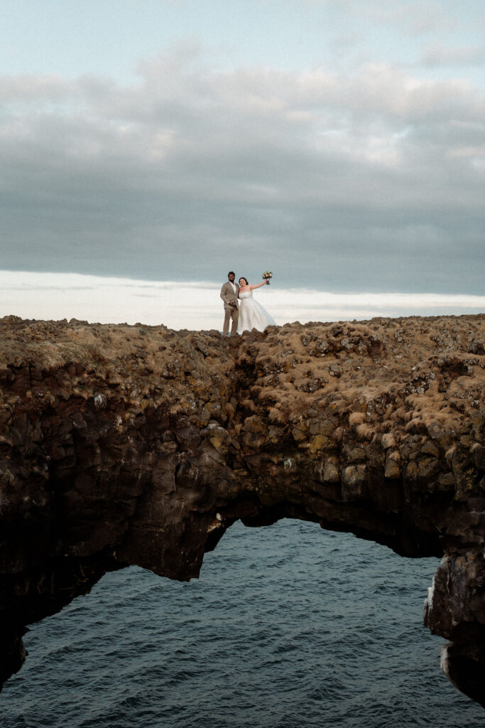 Bride and groom stand on rocky cliff above ocean, bride holding bouquet up. Overcast sky sets a dramatic, romantic scene at the best place to elope in iceland, snæfellsnes