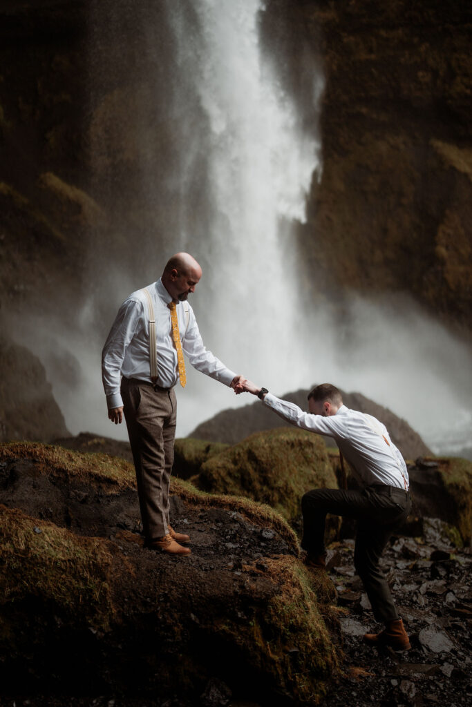 Two grooms, LGBTQ+ couple, one helping the other stand on rocks near a waterfall. The setting is lush and misty, evoking a bonding moment during iceland elopement