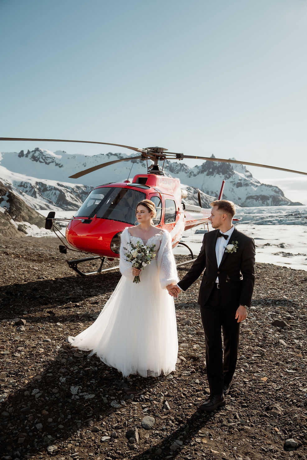 how much does it cost to elope in iceland? Bride and groom holding hands in front of a red helicopter on a rocky, snowy mountain landscape. Bride in white, groom in black suit.