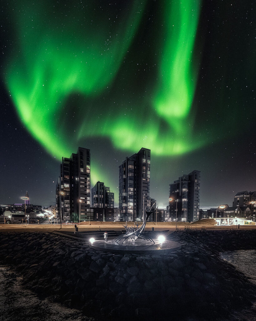 Northern lights in vivid green swirl over iceland cityscape with modern buildings. A metallic sculpture is illuminated in the foreground. 