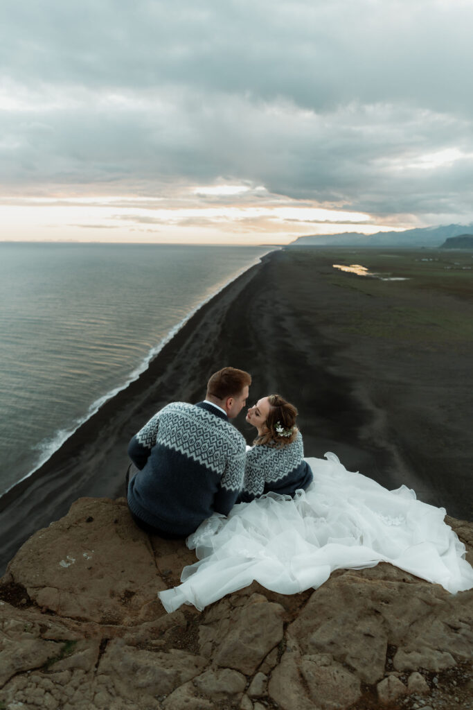 bride and groom sit, in sweaters, on a cliff edge, overlooking the black sand beach during adventure elopement in iceland.